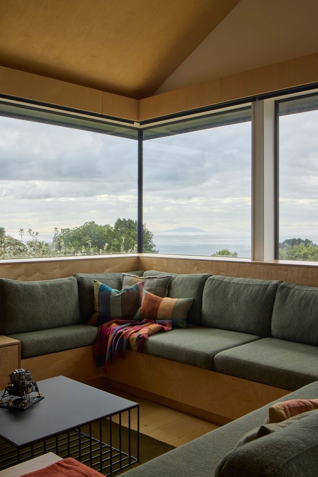 A cozy living room with a large built-in L-shaped sectional sofa wrapping around a corner of floor-to-ceiling glazing. Warm wood panelling covers the ceiling, window surrounds, and built-in seating base, while expansive corner windows overlook a cloudy coastal or lakeside landscape. A black metal coffee table, textured cushions, and a colourful throw add contrast to the muted green upholstery. The space has a refined mid-century-inspired character with strong indoor-outdoor connection.