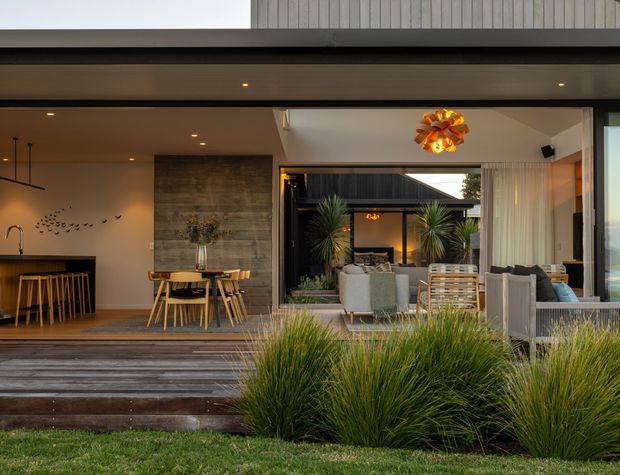 Contemporary indoor-outdoor living and dining area opening onto a landscaped lawn through wide glazed sliding doors. The space features dark timber decking, a concrete-look feature wall, timber dining furniture, a kitchen island, neutral upholstered outdoor-style seating, sheer curtains, recessed lighting, and a sculptural pendant. A planted courtyard or garden is visible beyond the main living zone.
