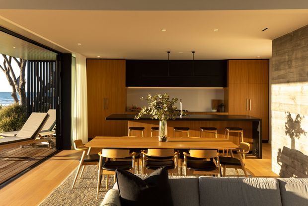 Warm, open-plan dining and kitchen interior with a timber dining table and chairs in the foreground, a contemporary black and timber kitchen with a long island behind it, and a grey sofa partially visible at the bottom. Full-height glazing opens to a timber deck and outdoor seating, while a textured concrete wall forms the right side. Recessed ceiling lights and pendant lights provide a refined, sunlit atmosphere.