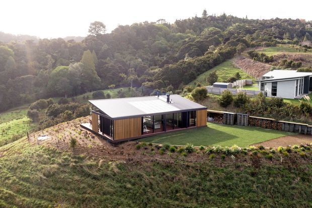 Aerial view of a modern single-storey home set into a steep, heavily landscaped hillside. The compact rectangular dwelling has a low-pitched metal roof, dark window and door frames, warm vertical timber cladding, and broad glazed openings facing a neatly clipped lawn. A second contemporary home is visible higher on the slope, surrounded by dense green vegetation and terraced garden areas.