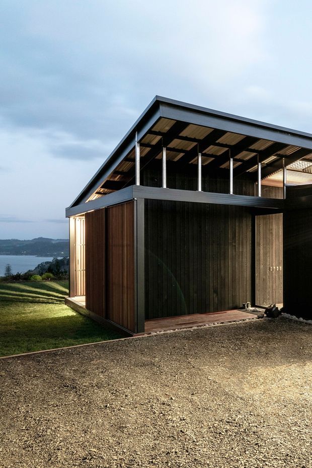 Exterior view of a contemporary dark-clad house on a coastal or lakeside hillside site. The building has a sharply pitched roof, a deep covered upper-level terrace or verandah with slender steel posts, dark vertical timber cladding, warm timber soffits, and large glazed openings. Gravel surrounds the foreground, with lawn and a body of water visible beyond the property.