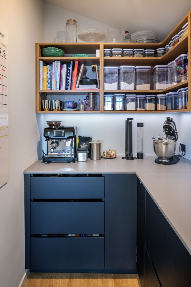 Compact, well-organised kitchen pantry and coffee station with dark navy built-in lower cabinetry, a pale stone or engineered-stone L-shaped benchtop, open timber shelving, and extensive clear storage containers. The shelves hold dry goods, books, crockery and glassware, while the benchtop contains an espresso machine, grinder, stand mixer and other coffee-making equipment. Warm timber flooring and under-shelf lighting add warmth to the otherwise dark-and-neutral fit-out.
