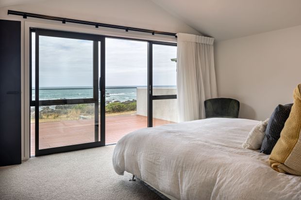 A bright coastal bedroom with a low-profile bed dressed in white linen, textured neutral cushions, and a dark upholstered armchair. Full-height black-framed sliding glass doors open to a timber deck and expansive ocean views, with white curtains and soft carpet creating a relaxed, airy atmosphere.