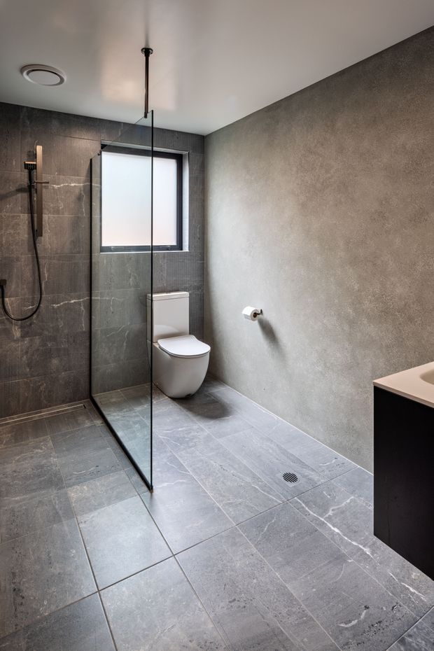A contemporary bathroom with a spacious open walk-in shower, frameless glass screen, wall-mounted rain and handheld shower fittings, toilet, and partial floating dark vanity. Large grey stone-look tiles cover the floor and shower walls, while the main wall has a smooth grey textured finish. A small frosted window provides natural light, and recessed ceiling lighting keeps the room evenly illuminated.