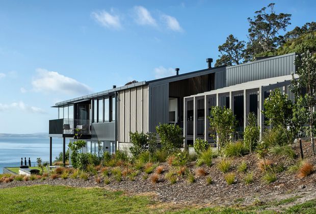 Contemporary multi-level coastal house set on a sloping, landscaped site overlooking a broad body of water. The home has a low-pitched roof, dark vertical metal cladding, extensive glazing, projecting balcony elements, and a partly enclosed outdoor terrace or verandah. Native-style planting surrounds the building.