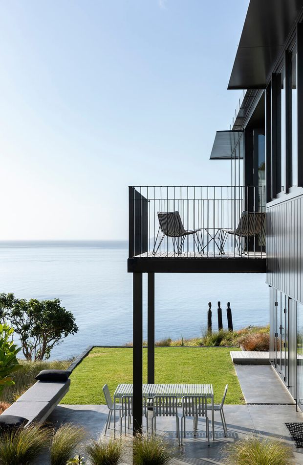 A contemporary modern coastal house viewed from its landscaped outdoor area, with a projecting elevated balcony overlooking calm water. The balcony has slim black metal railings, two lounge chairs and a small table, while the lower terrace contains outdoor dining furniture and a sun lounger. The building features dark metal cladding, extensive glazing, projecting flat roof planes and vertical supports. Grasses and clipped lawn frame the paved terrace and waterfront setting.