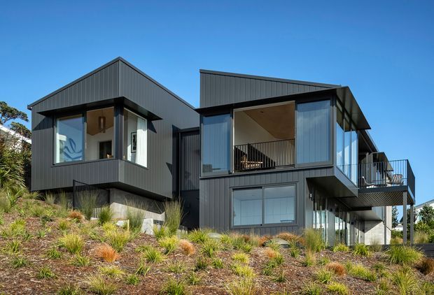 Exterior view of a contemporary multi-level house on a landscaped sloping site. The building has dark grey vertical metal cladding, angular projecting volumes, large glazed openings, balconies, and a mix of gabled and low-pitched roof forms. Native-style grasses and low planting cover the foreground beneath a clear blue sky.