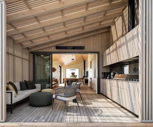 A light-filled enclosed outdoor living space or sunroom with timber slat walls and ceiling, timber decking, built-in outdoor kitchen cabinetry, and lounge seating. Large sliding glass doors open toward a connected interior living and dining area. Strong linear shadows from the slatted structure create a textured, contemporary atmosphere.