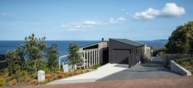 A contemporary dark-clad coastal residence viewed from the street, with a steeply sloping driveway and landscaped frontage. The building has a low-pitched asymmetrical roof, vertical cladding, large dark garage door, and a partially visible light-toned side structure. Ocean views are visible behind the house, with planted garden beds, timber fencing, and a white railing emphasizing the elevated sloping site.