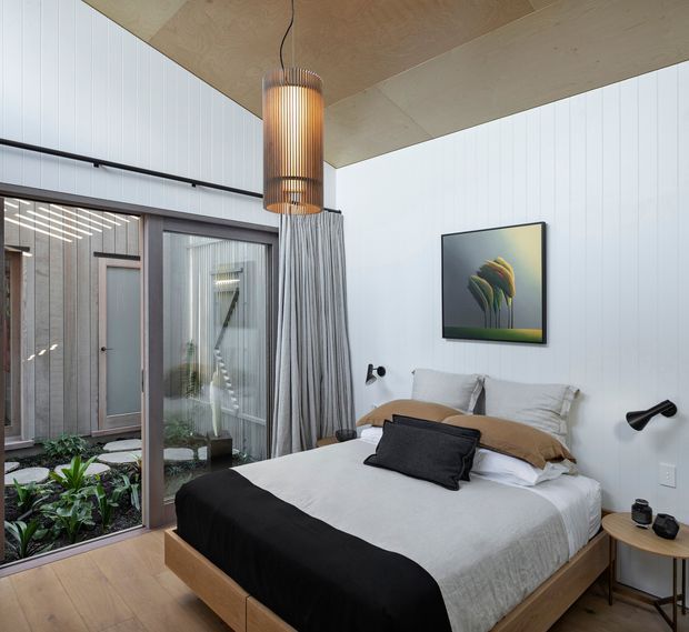 A contemporary bedroom with a low timber bed, layered neutral bedding, white vertical wall panelling, and a warm plywood ceiling. Full-height glazed doors open to a planted internal courtyard, with sheer curtains, black wall-mounted reading lights, a timber pendant, and restrained modern artwork.