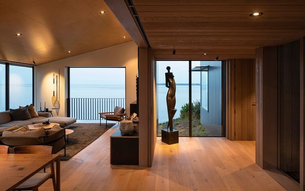 A warmly lit contemporary coastal interior showing an open-plan living room and entry hall. The living area has low-profile seating, a dark rug, timber furniture, and expansive glazing with ocean views beyond a balustrade. The adjacent circulation space features a timber-lined ceiling and walls, wide timber flooring, recessed downlights, full-height glass doors, and a sculptural display piece. The palette is predominantly warm timber, beige, brown, black, and blue from the view outside.
