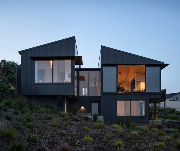 Exterior architectural photograph of a contemporary multi-level home on a sloping landscaped site. The dark charcoal vertical-clad facade is composed of offset box-like volumes with two asymmetrical pitched roof forms, expansive glazing, and a warm timber-lined recessed upper-level balcony or living space. Native-style planting and rock landscaping surround the house, with neighbouring homes visible in the background.