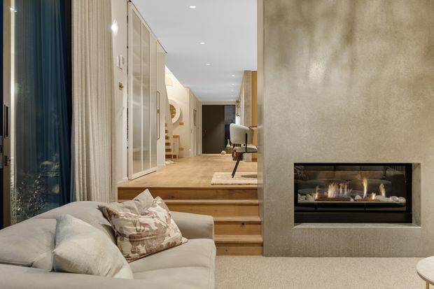 A refined contemporary living room with a light upholstered sofa in the foreground, a recessed linear fireplace set into a textured plaster feature wall, and warm timber steps leading to a raised hallway or study area. Floor-to-ceiling curtains and glazing line the left side, while built-in cabinetry and soft neutral finishes create a calm, luxurious interior.