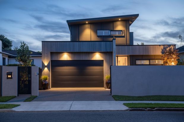 Contemporary two-storey urban residence photographed at dusk, featuring a flat-roofed upper volume, dark vertical cladding, warm integrated lighting, a large sectional garage door, boundary walls, a pedestrian gate, and a concrete driveway.