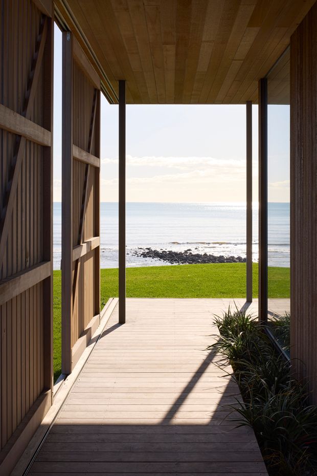 A timber-clad covered passage or entry opens directly onto a sunlit timber deck overlooking a calm ocean shoreline. Vertical timber screening, dark slender posts, and a timber-lined ceiling frame the coastal view, with low coastal planting along the right edge.