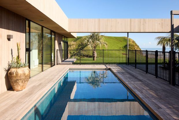 A contemporary architectural courtyard with a rectangular swimming pool surrounded by timber decking. The low-slung building has warm vertical timber cladding, extensive floor-to-ceiling glazing, and a projecting timber-lined soffit. Black metal balustrades frame views toward a grassy coastal headland and palm trees, creating a bright indoor-outdoor setting.