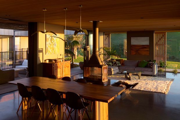Warm, open-plan mid-century-inspired living and dining space with extensive glazing, a timber-lined ceiling, polished dark flooring, and a central freestanding wood-burning fireplace. A large timber dining table with sculptural dark chairs occupies the foreground, while a low sofa seating area, built-in cabinetry, indoor plants, and a covered outdoor terrace are visible beyond.