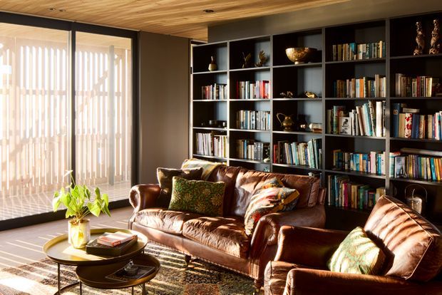 A warm, richly styled living room with a large dark built-in bookshelf spanning the rear wall, filled with books and decorative objects. A brown leather sofa and matching armchair sit around layered round coffee tables and a patterned rug. Full-height glazing with vertical blinds brings in strong sunlight, while the timber-lined ceiling adds warmth.