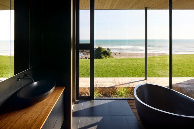 A contemporary bathroom opening directly onto a lawn and beachfront, with a freestanding dark bathtub, black vessel basin, timber vanity, dark wall and floor finishes, and extensive floor-to-ceiling glazing. The ocean and sandy beach are clearly visible beyond the landscaped lawn.