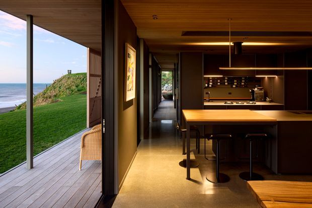 A sophisticated open-plan kitchen and living area opening directly onto a timber deck overlooking the ocean. The interior features dark built-in cabinetry, a warm timber island and ceiling, integrated lighting, a dark tiled or stone splashback, and contemporary bar stools. Full-height glazing and sliding doors connect the kitchen to the coastal outdoor setting.