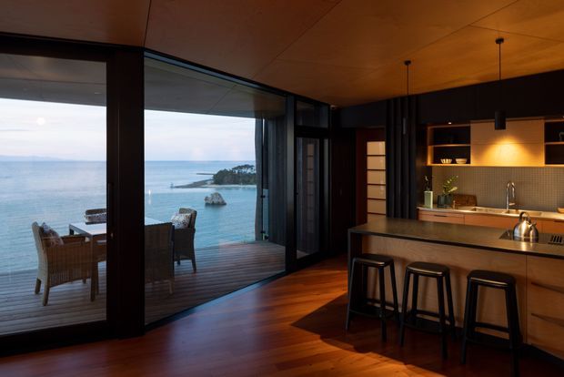 A moody, contemporary open-plan kitchen opening onto a timber deck with expansive floor-to-ceiling glazing and a visible coastal waterside outlook. The kitchen features warm timber cabinetry and ceiling panels, a dark textured tiled splashback, an island with a light stone benchtop and three stools, open shelving with integrated lighting, black pendant lights, and dark-framed sliding glass doors.