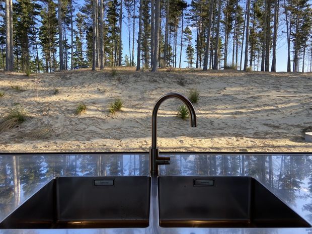 Close-up view of a contemporary kitchen sink area with a large dark double-bowl undermount sink and curved dark metal gooseneck tap. A highly reflective splashback or benchtop surface mirrors the surrounding pine trees, while a sandy, tree-covered landscape is visible through the window behind the sink.
