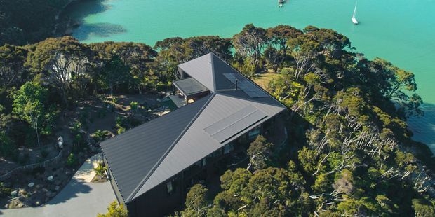 Aerial view of a dark-clad, multi-wing house nestled among dense coastal trees on a steep waterfront site. The building has intersecting pitched roofs with dark metal roofing, skylights, and expansive glazing, with turquoise water and boats visible beyond the property.