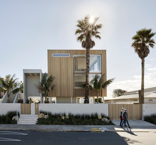Street-level exterior view of a contemporary two-storey residence with a box-like form, vertical timber cladding, white rendered walls, a projecting upper floor, and large glazed openings screened by vertical timber battens. Palm trees, low coastal-style landscaping, a pedestrian sidewalk, and a driveway are visible in front of the property.