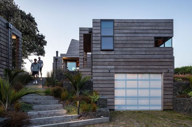 Exterior view of a contemporary multi-level home on a landscaped sloping site. The building has dark horizontal timber-like cladding, strong rectilinear volumes, large glazed openings, and an integrated garage with a translucent paneled door. Stepped concrete paths, native-style planting, gabion retaining walls, and a small group of people are visible.