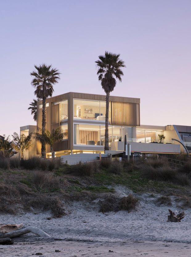A contemporary multi-level beachfront residence photographed at dusk, with expansive glazing, warm interior lighting, vertical timber screening, clean rectilinear volumes, and a flat roofline. Palm trees and sandy coastal vegetation frame the house, with the beach in the foreground.