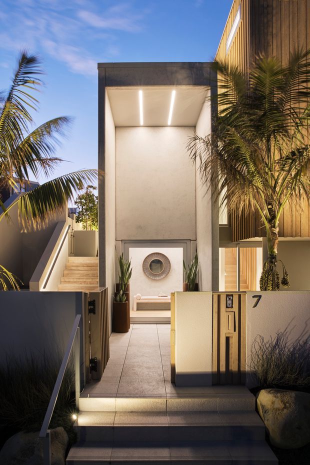A dusk exterior view of a contemporary residential entry. Stepped tiled stairs lead through an open gate to a covered entrance courtyard, with a tall recessed portico illuminated by linear ceiling lights. The entry includes a built-in bench, circular wall feature, potted plants, palms, a timber-clad upper facade, rendered walls, and a visible house number 7.