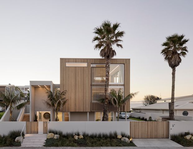 Contemporary multi-level residential facade with vertical timber cladding, concrete elements, screened glazing, and a recessed entry. Tall palm trees, landscaped planting, a timber privacy gate, and neighbouring homes frame the coastal suburban streetscape.