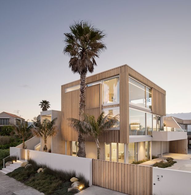 Contemporary multi-storey residential house with a strong timber-clad cubic form, extensive floor-to-ceiling glazing, recessed balconies, and vertical timber screening. The landscaped frontage includes palm trees, low planting, stone accents, and a rendered boundary wall with timber gates. A neighbouring suburban coastal-style setting is visible in the background.