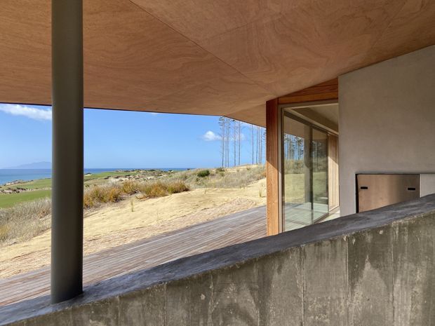 A sheltered exterior terrace or covered outdoor living area overlooking a coastal landscape. The space features a broad plywood-lined ceiling, a dark cylindrical structural post, a weathered concrete parapet wall, timber decking, timber-framed glazing and a built-in concrete or masonry fireplace with a simple dark opening. Dunes, grasses and the ocean are visible beyond, giving the image a restrained, contemporary coastal architectural character.