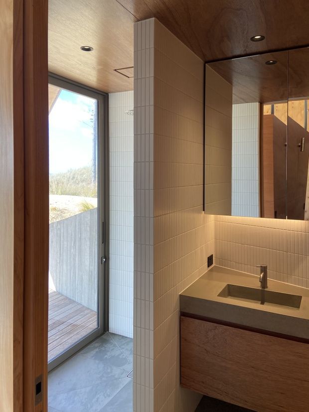 A compact, contemporary bathroom vanity area with a wall-mounted timber vanity, integrated rectangular basin, slim metal tapware, and a large illuminated mirror. Pale small-format grid tiles wrap the walls and partition, while the ceiling and surrounding door frames use warm timber. A glazed door opens to a timber deck and exterior landscape.