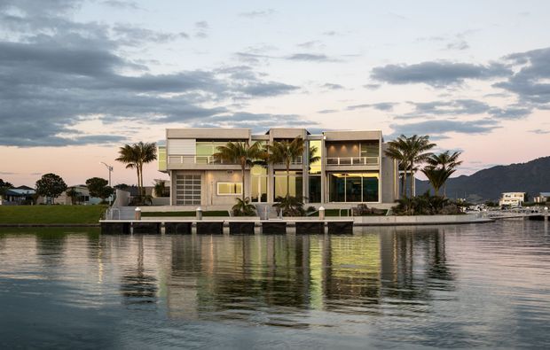 A modern multi-storey waterfront residence viewed across calm water at dusk. The house has a rectilinear composition with extensive glazing, projecting box-like upper levels, pale rendered walls, dark window frames, a flat roofline, and a landscaped waterfront edge with palms and a small dock. Distant hills and neighbouring waterfront buildings are visible in the background.