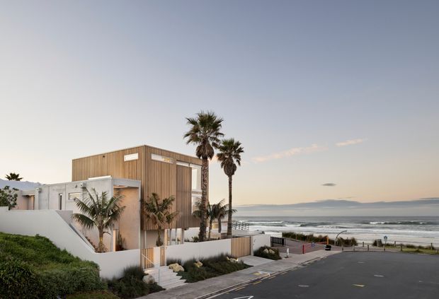 Contemporary multi-level coastal residence with a boxy timber-clad upper volume, white rendered walls, and large glazed openings. The house sits beside a beachfront road, with palm trees, landscaped planting, and ocean waves visible beyond.