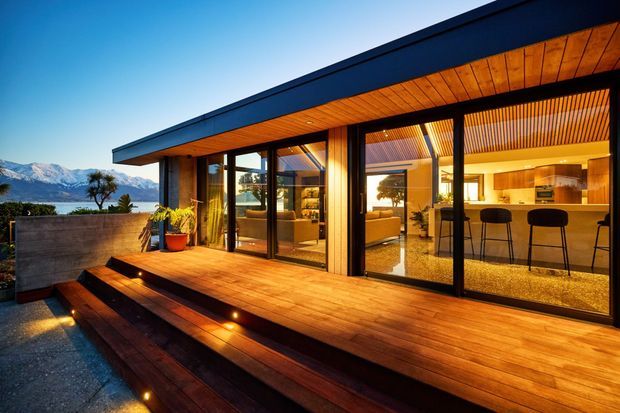 Modern single-storey house exterior at dusk, featuring a broad timber-clad deck with illuminated steps, extensive floor-to-ceiling glazing, dark metal framing, and a warm timber soffit. A contemporary open-plan living and kitchen area is visible through the glass, with a kitchen island and bar seating. Mountains and water are visible in the distant background.