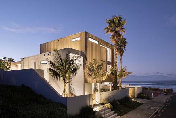 A contemporary multi-level coastal residence with a boxy form, white rendered walls, and a prominent upper volume clad in vertical timber battens. Tall narrow windows and exterior stairways frame the house, which sits beside a descending residential street overlooking the ocean. Palm trees and low boundary walls reinforce the coastal setting, with the building illuminated in warm evening light beneath a clear blue sky.