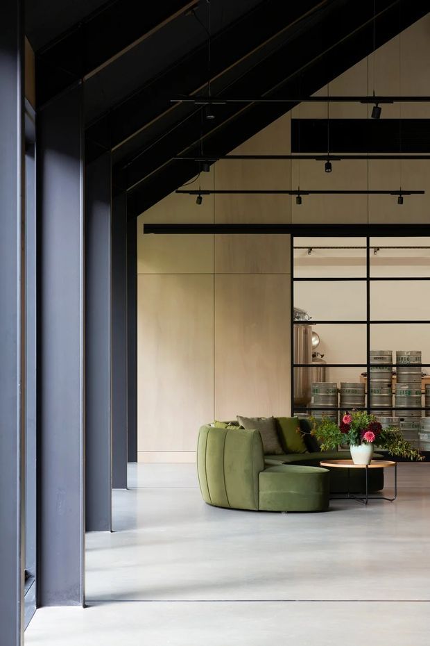 A sophisticated open-plan interior with a dark exposed structural ceiling, black steel-framed glazing and partitions, pale timber wall panelling, and a polished concrete floor. A curved olive-green lounge chair and matching cushions sit beside a low round table with flowers, while shelving displays cylindrical objects behind the seating area. The space has a high industrial-style ceiling, strong linear detailing, and a refined, muted palette.