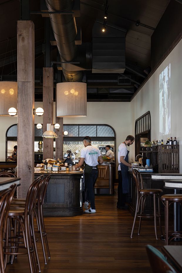 A vertically framed photograph of a stylish restaurant interior with a bar and service counter, dark timber flooring, exposed black industrial ceiling services and ductwork, arched glazed windows, timber columns, pendant lighting, and high stools. Staff are working behind the counter, with tables and seating arranged throughout the dining space. The atmosphere is warm, dark, and refined with a strong industrial character.