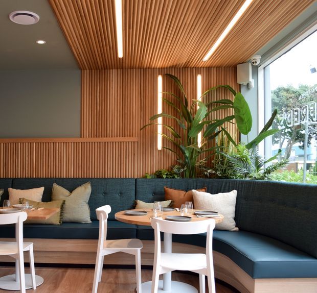 A bright contemporary restaurant or hospitality dining area with curved built-in dark teal banquette seating, small round timber tables, white chairs, and neutral cushions. The space features extensive vertical timber slat wall and ceiling lining, integrated linear lighting, indoor tropical planting, and large glazing overlooking greenery outside.