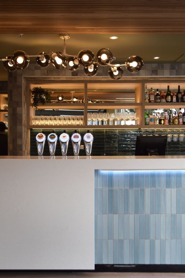Interior photograph of a refined commercial bar, likely a restaurant or hospitality venue. The bar features a pale solid counter, blue vertical tile wall, dark glossy backsplash tile, timber-framed open shelving with bottles and glassware, beer taps, and a sculptural cluster of round pendant lights beneath a timber slatted ceiling. The palette is warm timber, charcoal, cream, and cool blue tile.