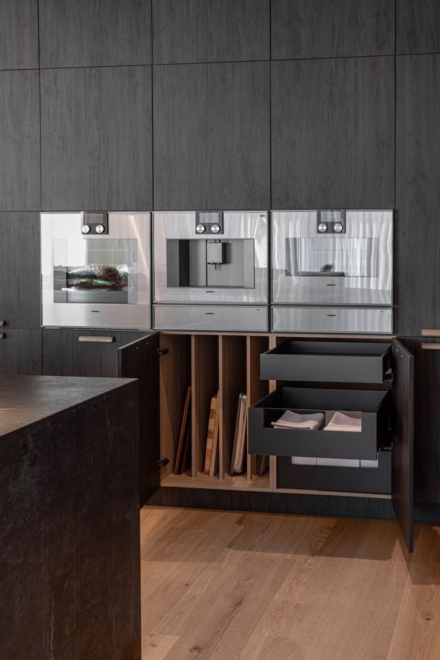 Close-up of a sophisticated dark timber kitchen wall with integrated stainless-steel ovens and coffee appliance, extensive cabinetry, open pull-out drawers, vertical storage for trays and boards, and a dark stone island benchtop. Warm timber flooring contrasts with the charcoal cabinetry and brushed metal appliances.