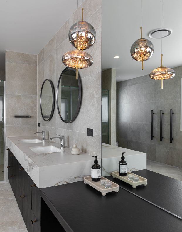 Contemporary bathroom vanity with a large white marble-look double basin benchtop, dark timber cabinetry, grey large-format wall and floor tiles, twin oval black-framed mirrors, and sculptural spherical pendant lights. A full-height mirror reflects the tiled shower area with black hardware and handles.