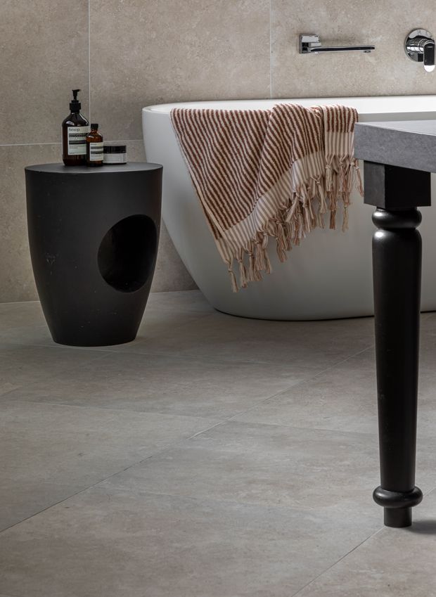 A refined contemporary bathroom featuring a freestanding white oval bathtub, large-format grey-beige stone-look wall and floor tiles, a black sculptural side table, and a black vanity leg with a grey benchtop. Chrome wall-mounted tapware and a striped neutral towel add functional and decorative detail.