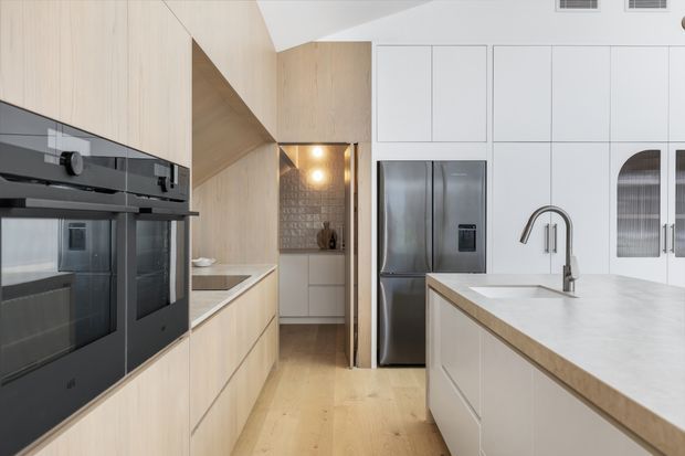 Bright contemporary kitchen with pale timber cabinetry, integrated black ovens, a large white island with a stone benchtop, stainless-steel refrigerator, and a textured tiled splashback in a recessed preparation area. The space has clean-lined joinery, light timber flooring, and understated modern fixtures.