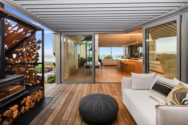 A refined covered outdoor terrace or balcony overlooking a coastal setting, with timber decking, a white outdoor sofa, black woven ottoman, sculptural firewood storage and decorative metal screen. Large sliding glass doors open into a warm contemporary kitchen and dining area, with timber cabinetry, a central island and bar seating. A louvred overhead roof provides shade while maintaining an open-air feel.