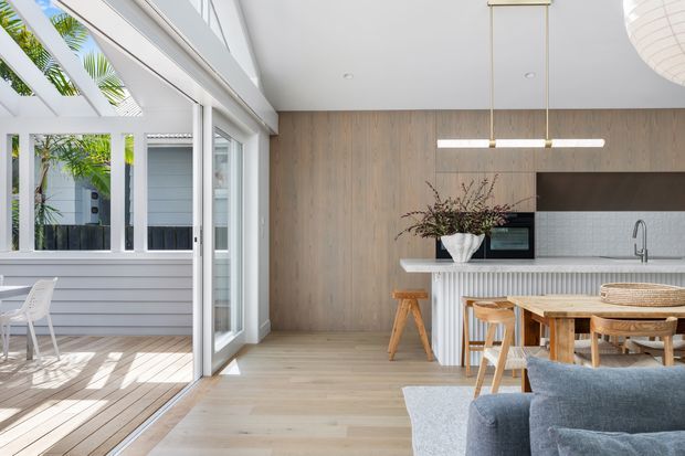 Bright open-plan interior combining a contemporary kitchen, dining area and living space, with a large glazed opening to a light-filled covered outdoor terrace. The kitchen features a warm vertical timber wall, white fluted island cabinetry, dark splashback cabinetry, pale timber flooring, and a linear pendant light. A timber dining table and chairs sit beside the island, while a blue-grey sofa occupies the foreground. The space has a relaxed, coastal-influenced contemporary feel with strong indoor-outdoor connection.