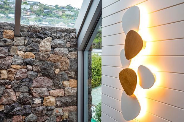 A contemporary interior feature wall beside a tall angled glazed opening. The wall combines horizontal white wall panelling with an irregular stacked-stone masonry section, and features sculptural oval wall lights or decorative panels in white and warm timber tones. Through the glazing, a hillside residential landscape and water are visible.